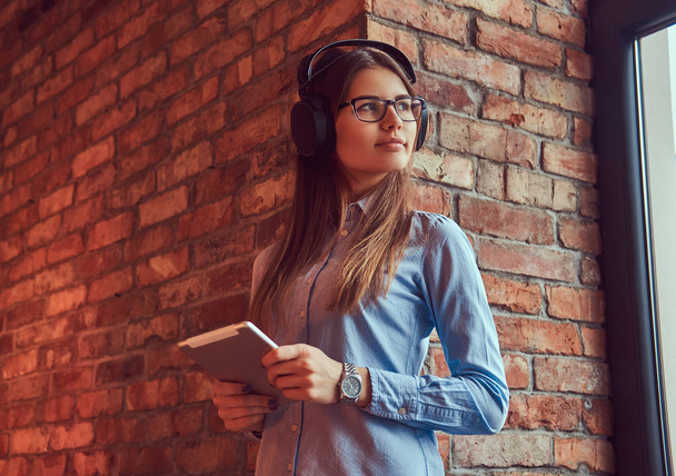 Μια νεαρή γυναίκα μελαχρινή σε γυαλιά και μπλε πουκάμισο. Στέκεται ενάντια σε έναν τοίχο από τούβλα και να ακούτε μουσική στα ακουστικά με το tablet. - Φωτογραφία, εικόνα