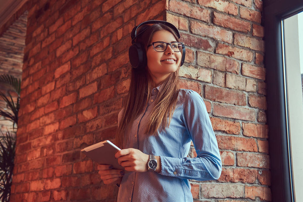 A young brunette woman in glasses and blue shirt. Smiling and standing against a brick wall and listening to music on headphones with the tablet. - Foto, Imagen