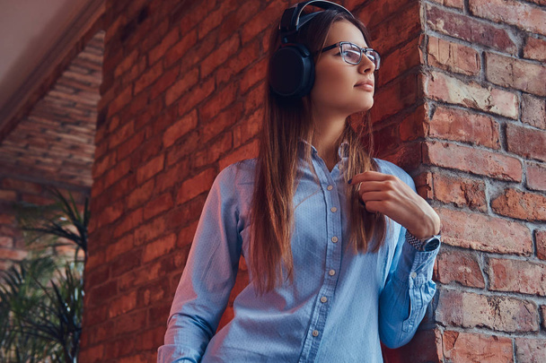 Retrato de una atractiva morena encantadora en gafas y camisa azul escuchando música en auriculares apoyados en una pared de ladrillo en una habitación con diseño loft
. - Foto, imagen