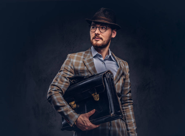 A bearded man wearing casual suit in a hat and glasses holds suitcase - Fotoğraf, Görsel