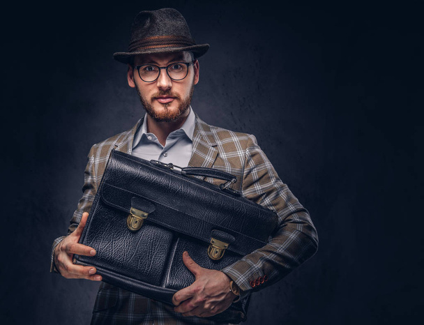 Een bebaarde man dragen casual pak in een hoed en bril houdt koffer - Foto, afbeelding