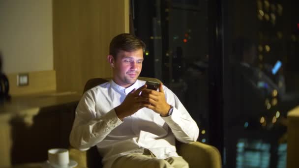 Young, beautiful man using a smartphone, on a chair in a room with a panoramic window overlooking the skyscrapers at night. 4k, blur the background. - Záběry, video