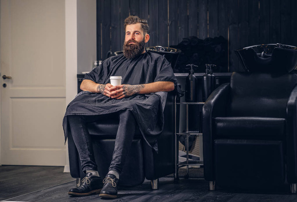 Przystojny, brodaty mężczyzna z tatuażem na jego ramionach pije kawę przed mycia włosów w salonie Fryzjer. - Zdjęcie, obraz