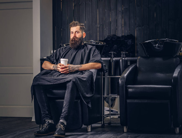 Handsome bearded man with a tattoo on his arms drinks coffee before hair wash in a hairdressers salon. - Photo, Image