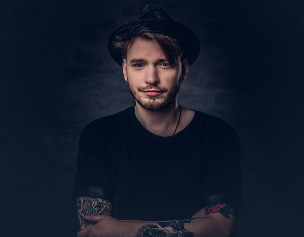 Portrait of handsome bearded hipster with crossed tattooed arms, dressed in a black t-shirt and hat. Isolated on a dark background. - Zdjęcie, obraz