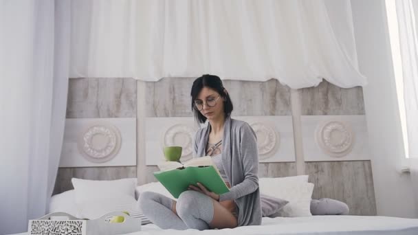 A charming girl in glasses sitting in bed, drinking coffee and read book at home - Metraje, vídeo