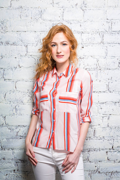 A beautiful young woman student with red curly hair and freckles is leaning against a brick wall of gray color. Dressed in a red striped shirt and white pants - Φωτογραφία, εικόνα