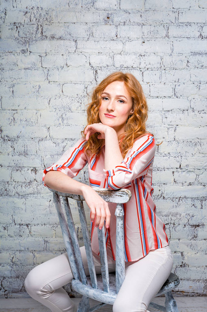 portrait Beautiful young woman student with red curly hair and freckles on her face sitting on a wooden chair on a brick wall background in gray. Dressed in a red striped shirt - Foto, afbeelding