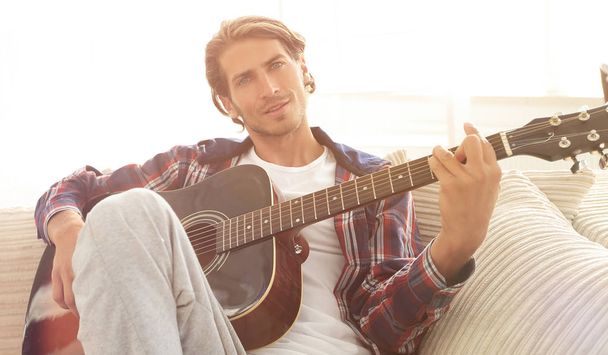 modern guy with guitar sitting on sofa in living room. - 写真・画像