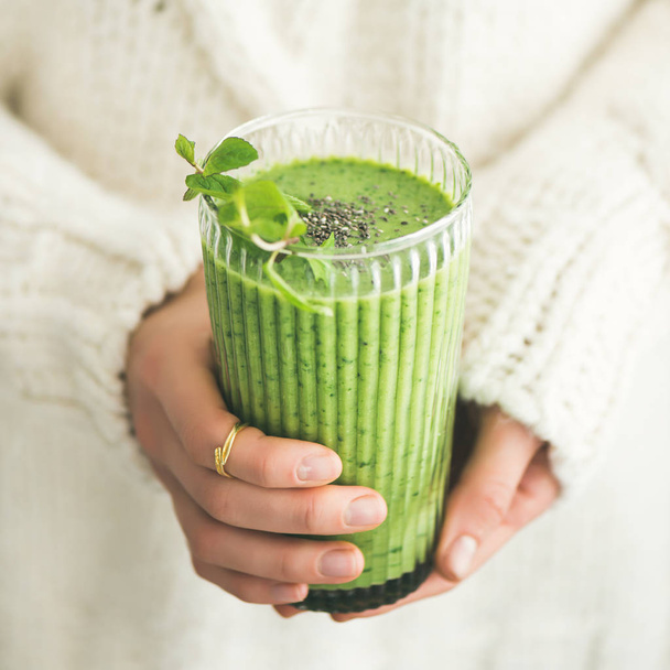 Matcha green vegan smoothie with chia seeds and mint in glass in hands of female wearing white sweater, square crop. Clean eating, detox, alkaline diet, weight loss concept - 写真・画像