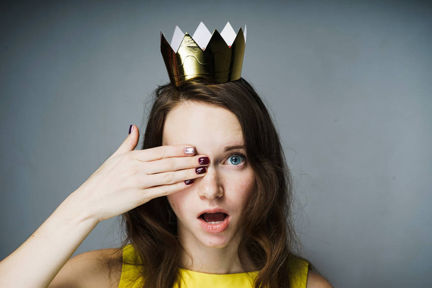 a surprised young girl in a yellow dress closed one eye with her hand, a golden crown on her head - Photo, Image