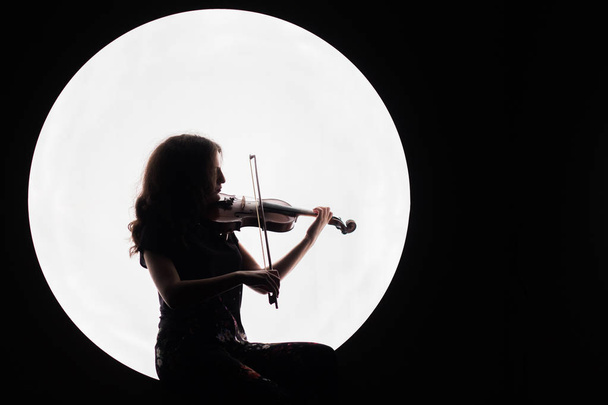 Silhouet van een prachtige brunette meisje viool spelen. Concept voor muzieknieuws. Kopieer ruimte. Witte cirkel als de maan op een zwarte achtergrond. - Foto, afbeelding
