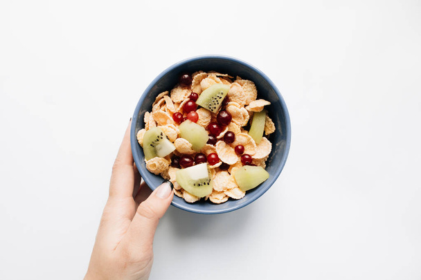 tazón de mano femenino con deliciosos copos de maíz crujientes, trozos de kiwi y arándanos sobre fondo blanco, primer plano, desayuno saludable
  - Foto, Imagen