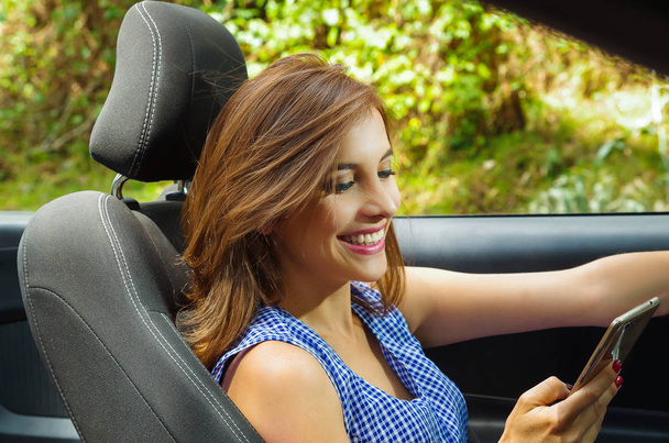 Close up of beautiful caucasian woman using her cellphone inside of the black car, while she is driving in a blurred nature background - Фото, изображение