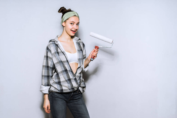 happy young girl in a large checkered shirt holds a roller for painting the walls, repair in the apartment - Foto, Bild
