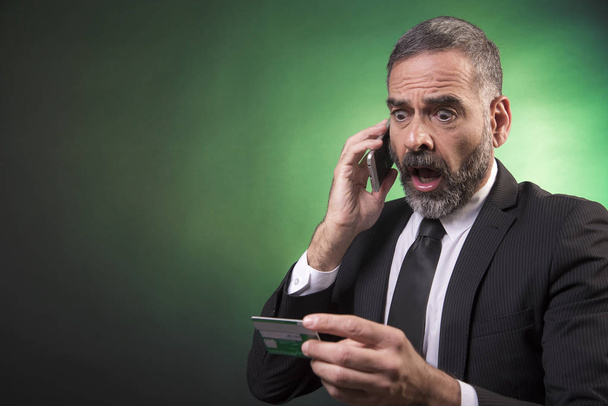 Homem de negócios sênior totalmente chocado com seu saldo de cartão de crédito, discutindo com o funcionário do banco sobre seu telefone celular
 - Foto, Imagem