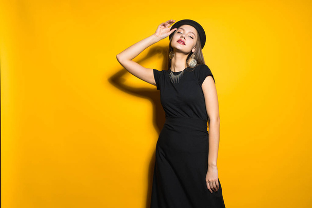 luxurious elegant young girl in black dress and hat posing on yellow background, demonstrating expensive jewelry - Photo, Image