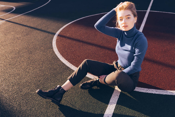 beautiful stylish young blond model girl in blue sweater sits on a sports field in the sun - Фото, зображення