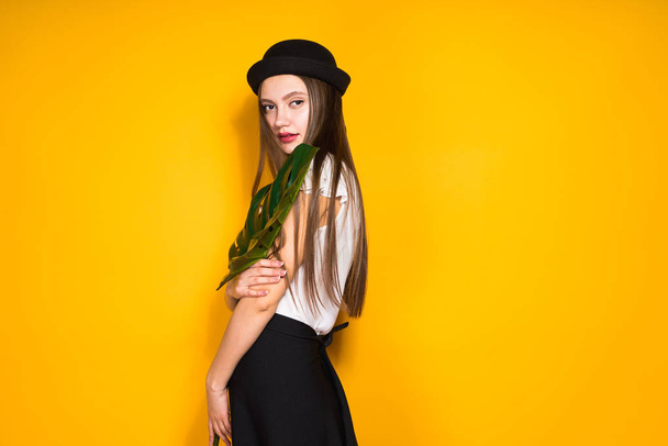 confident attractive girl in fashionable black hat posing on yellow background, holding a green leaf - Fotografie, Obrázek