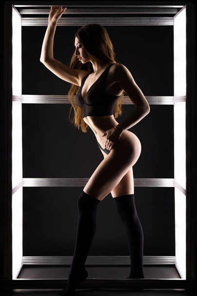 Young dark-haired woman in black  lingerie posing against white lights in black studio - Foto, afbeelding