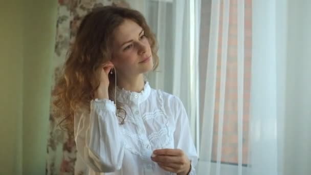 girl curly hair, listening to music in the headphones at the window, dancing, singing. portrait - Materiał filmowy, wideo