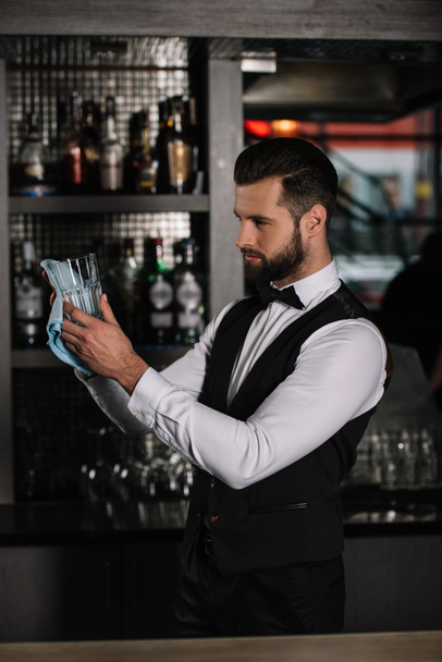 side view of handsome bartender cleaning glass with rag in evening - Фото, изображение