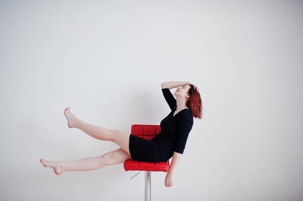 Red haired girl on black dress tunic sitting on red chair agains - Foto, imagen