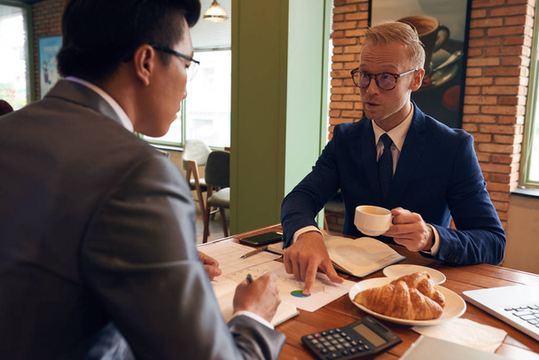 Businessman explaining diagram to his Vietnamese colleague during meeting in cafe - Photo, Image