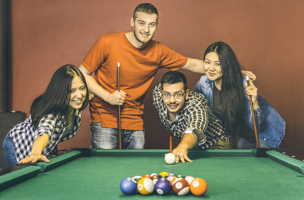 Young friends playing pool at billiard table saloon - Happy friendship concept with fashion people having fun together and sharing time at snooker gameroom - Warm vintage retro contrast filter - Photo, Image