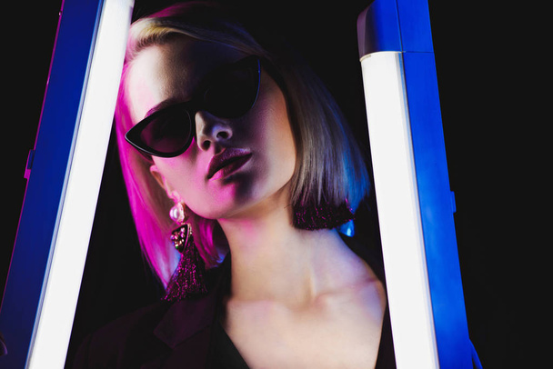 stylish attractive girl posing in sunglasses with two ultra violet lamps for fashion shoot, isolated on black - Foto, immagini
