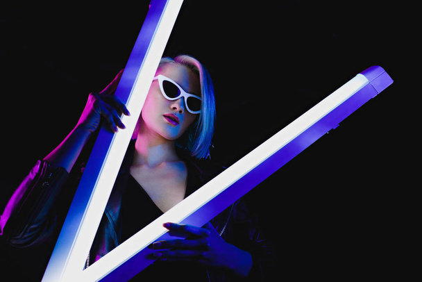 beautiful stylish model in sunglasses posing with two ultraviolet lamps for fashion shoot, isolated on black - Fotografie, Obrázek