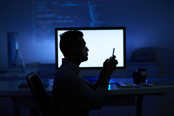 Ohjelmoija lukee tietoja älypuhelimella pimeässä toimistossa
 - Valokuva, kuva