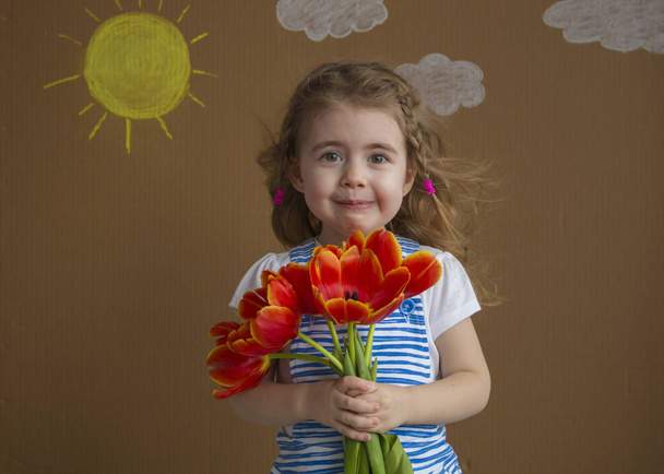 Emocionální portrét krásná dívka v modrých šatech s květy tulipánů v rukou na světlém pozadí s malovanými slunce a mraky - Fotografie, Obrázek