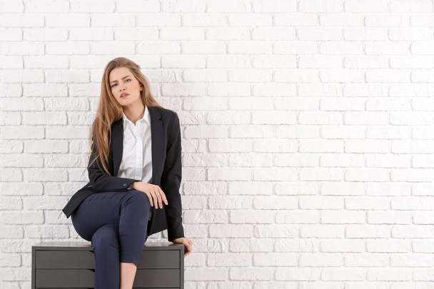 Beautiful fashionable woman in elegant suit sitting on chest of drawers against white brick wall - Foto, afbeelding
