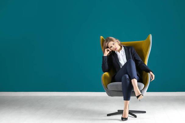 Beautiful fashionable woman in elegant suit sitting in armchair against color wall - Foto, imagen