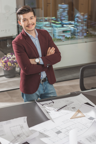 sonriente joven arquitecto con brazos cruzados cerca del lugar de trabajo en la oficina
 - Foto, imagen