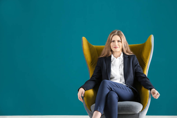 Beautiful fashionable woman in elegant suit sitting in armchair against color background - Foto, afbeelding