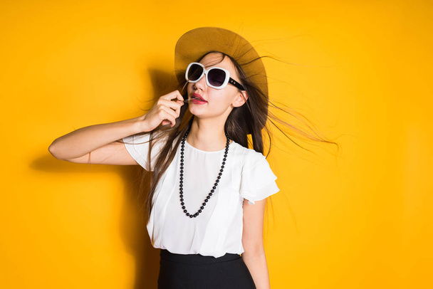 attractive young girl in fashion hat and black sunglasses posing on yellow background, hair flapping - Foto, immagini