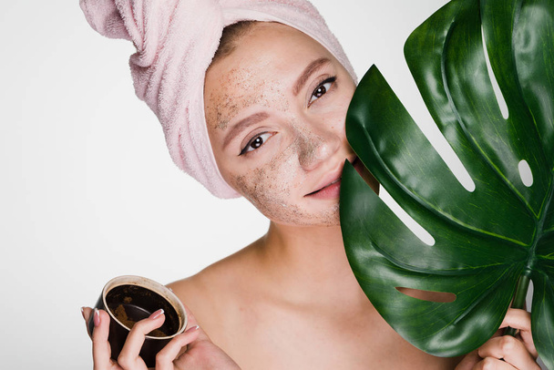 beautiful young girl with a pink towel on her head puts a coffee scrub on her face, holds a green leaf, day spa - Foto, Imagem