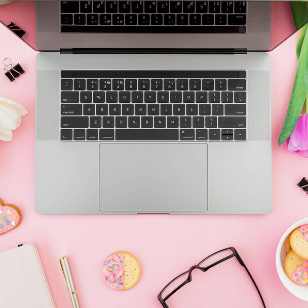 Top view of laptop surrounded by fresh tulips, sprinkled cookies and stationery on pink background - Photo, image
