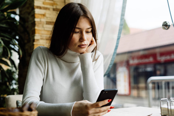 beautiful dark-haired girl in a white sweater sits in a cafe and looks in a smartphone - Foto, Imagen