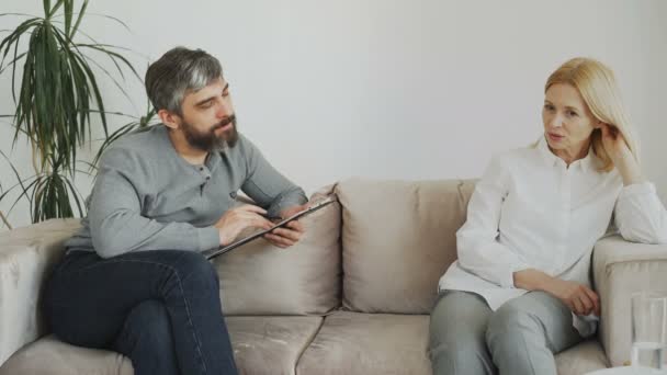 Psicólogo experimentado masculino que habla y escucha al paciente molesto y escribe notas en el portapapeles en su oficina
 - Imágenes, Vídeo