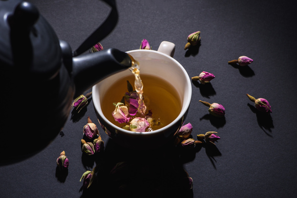 Tee aus schwarzem Wasserkocher in Tasse gießen und rosa Rosenknospen auf schwarz trocknen - Foto, Bild
