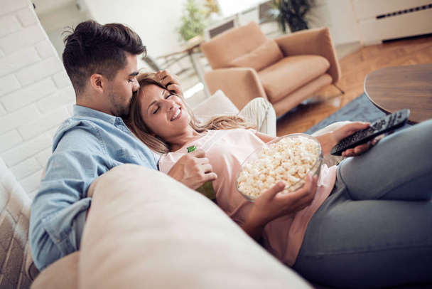 Casal jovem relaxando em seu apartamento, deitado no sofá, assistindo a um filme e comendo pipocas
. - Foto, Imagem