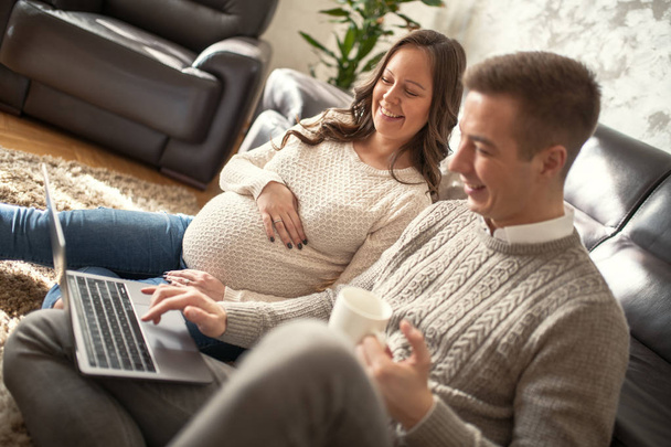 Χαρούμενο ζευγάρι χρησιμοποιώντας φορητό υπολογιστή στο σπίτι, νεαρή γυναίκα είναι έγκυος. - Φωτογραφία, εικόνα