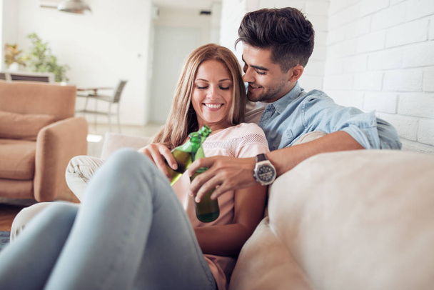 Giovane coppia seduta sul divano in soggiorno, con birra in mano e sorridente
. - Foto, immagini