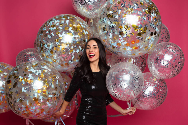 Stylish young smiling beautiful woman in black evening dress on a pink background with large multi-colored helium balls. Birthday party. Concept of gifts and congratulations. - 写真・画像