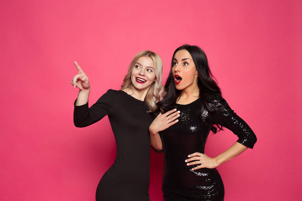 Look! Two beautiful smiling modern women in little black dresses posing in front of the camera on a pink background. - Foto, Imagem