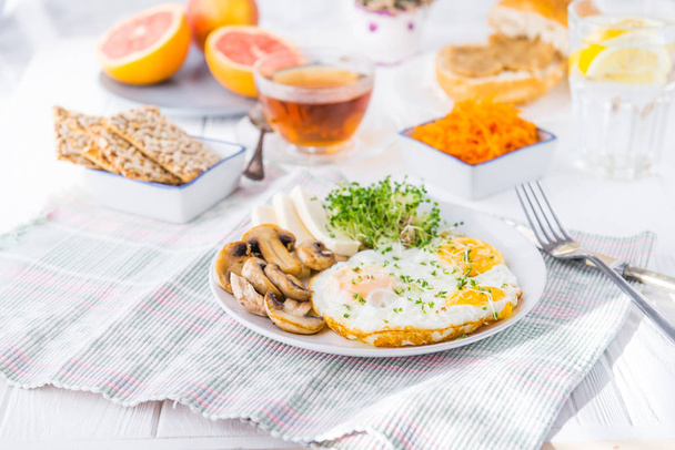 Healthy breakfast plate with scrambled eggs, cheese, grilled mushrooms and sprout micro greens and other snacks and drinks on the served white wooden table. Selective focus. - 写真・画像