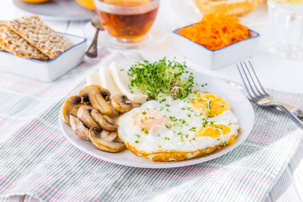 Healthy breakfast plate with scrambled eggs, cheese, grilled mushrooms and sprout micro greens and other snacks and drinks on the served white wooden table. Selective focus. - 写真・画像